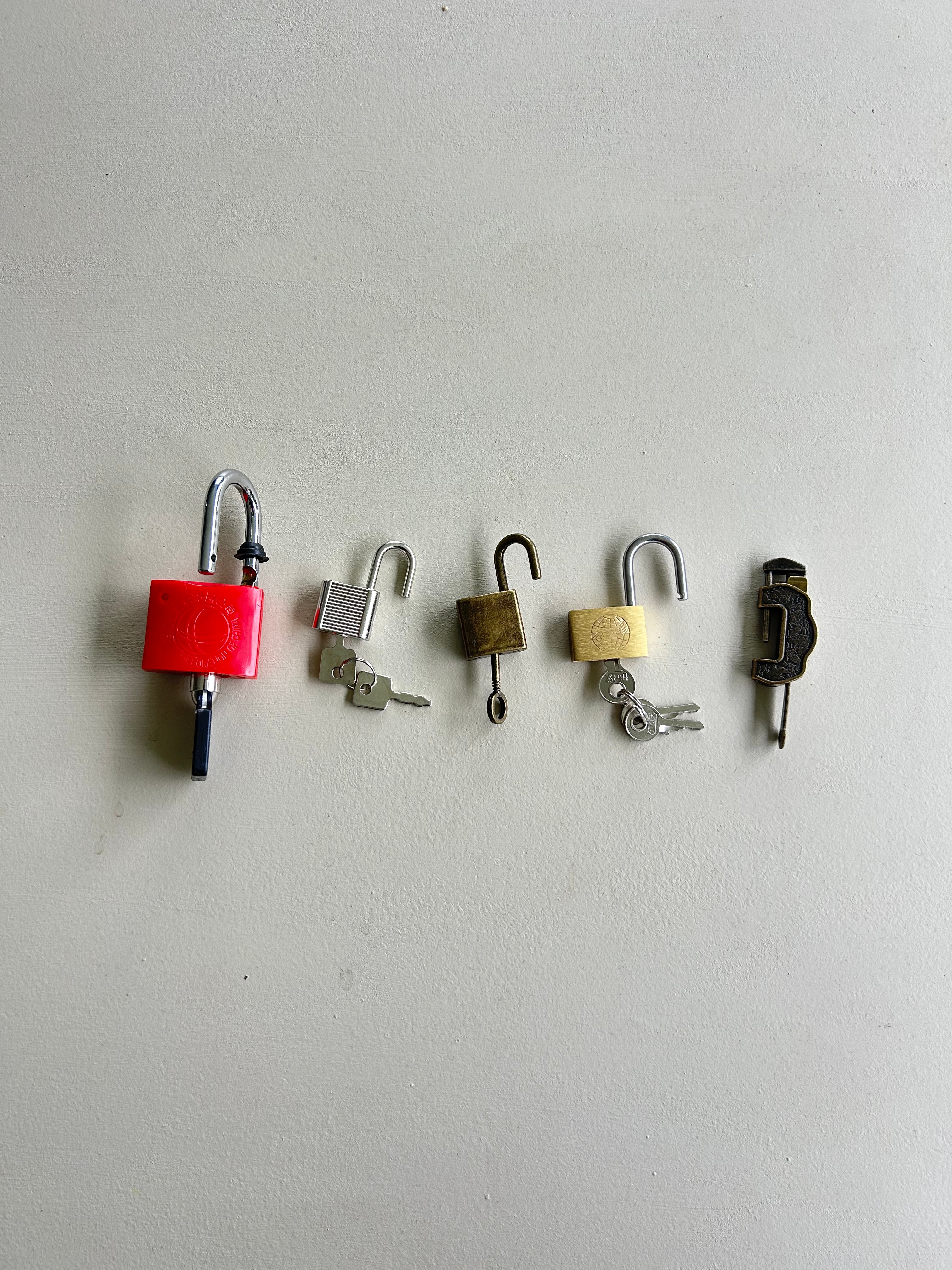 Five different padlocks and keys arranged in a row against a plain off-white wall. The locks include a bright red cylindrical padlock, a silver metal padlock with a key, a small bronze padlock, a cream padlock with a key, and a dark metal padlock. montessori