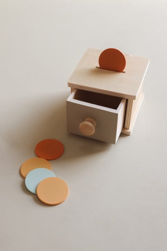 A minimalist wooden drawer toy with a partially open drawer and a small orange circular piece on top, shown on a warm off-white background with three loose matching pieces in orange, yellow, and light blue.