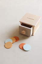 A wooden children's money box with an open drawer and a slot on the lid, shown on a warm beige background with several pastel-colored round coins scattered in front.