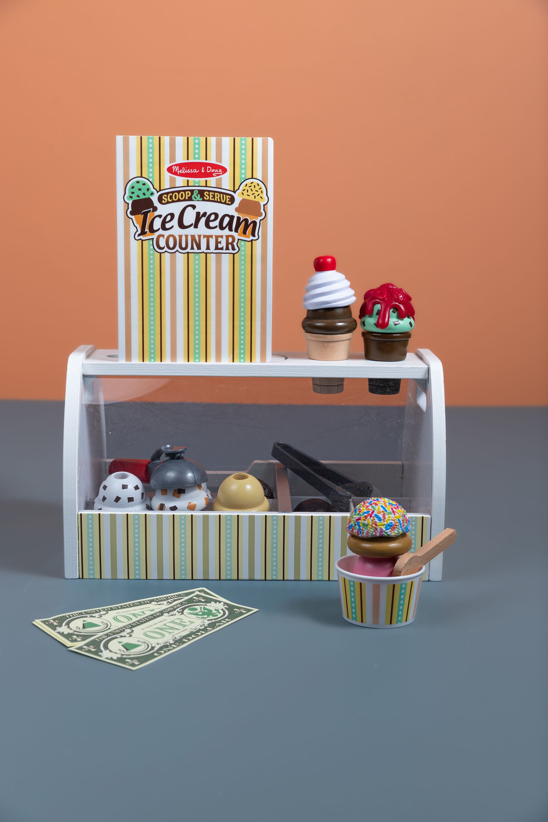 Melissa & Doug Scoop & Serve Ice Cream Counter wooden playset displayed with all 28 pieces including scoops, toppings, cones, cup, scooper, tongs, spoon, menu card, and pretend dollar bills arranged in front of the wooden counter