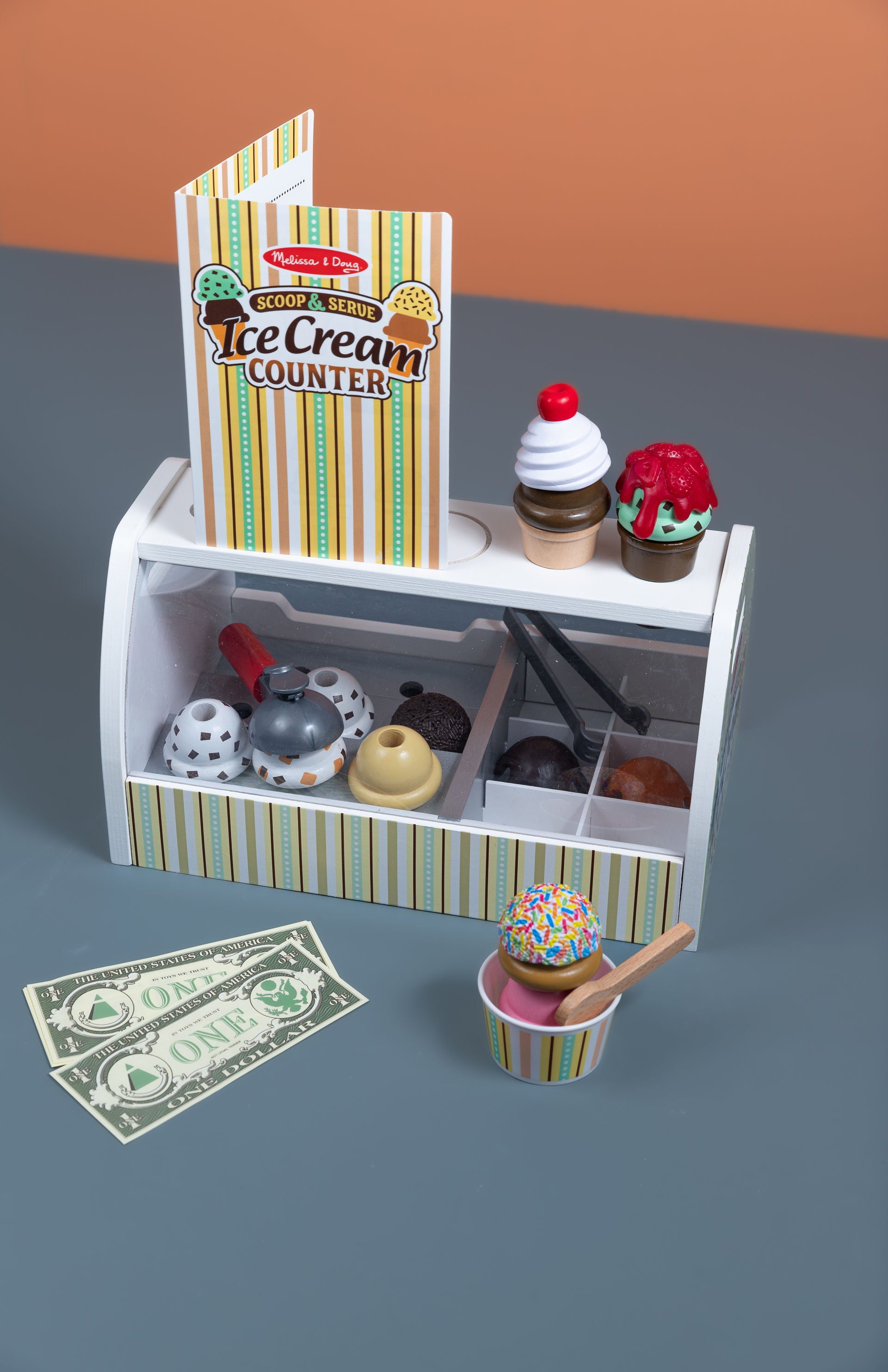 Toy ice cream counter set with ice cream cones, toppings, and money on a gray surface.