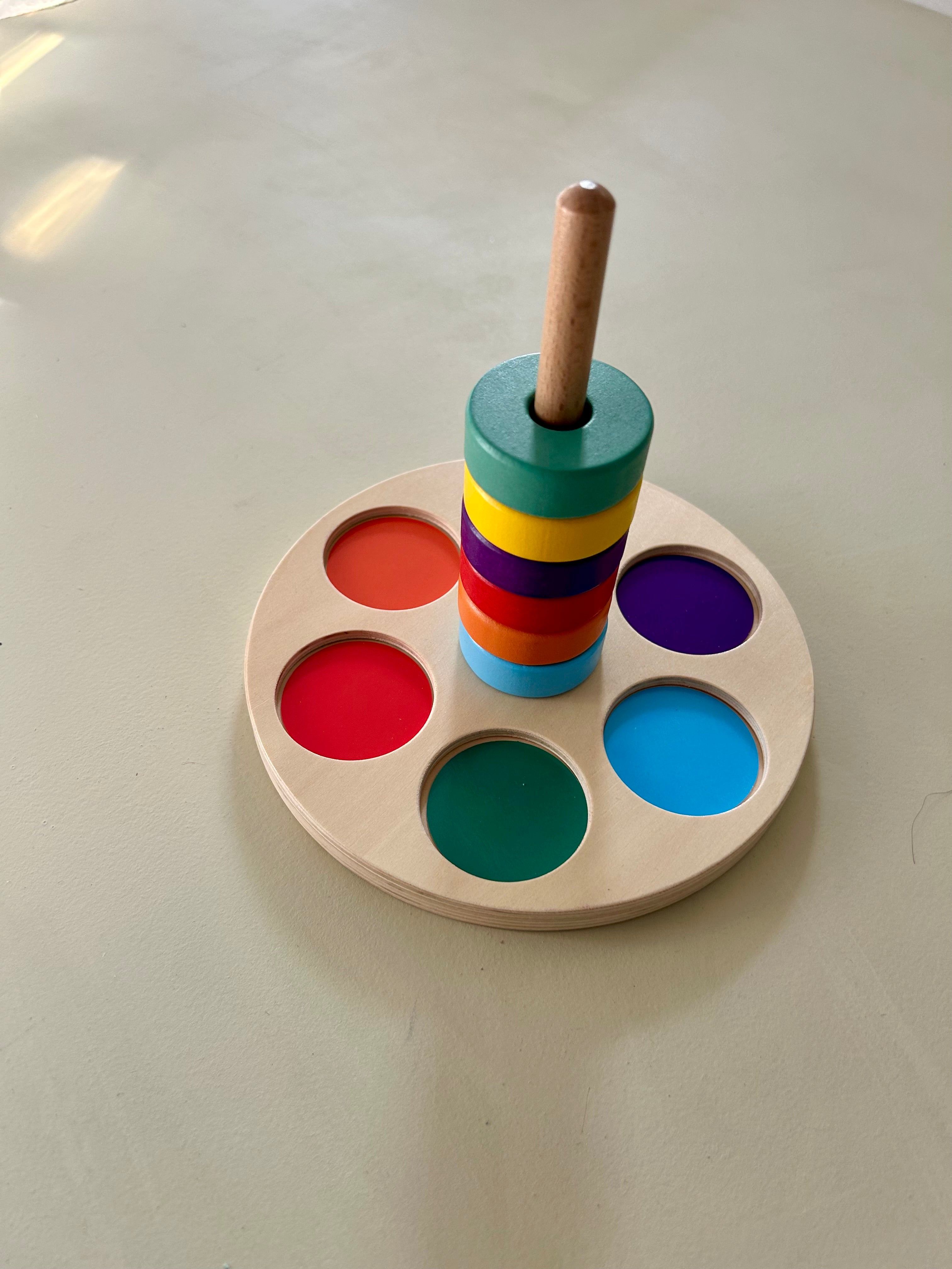 Wooden colour matching stacker on a pale sage surface. Six painted wooden rings are stacked on a central wooden peg: light blue, orange, red, purple, yellow and green. They sit on a round natural plywood base with recessed circles in orange, red, green, light blue and purple.