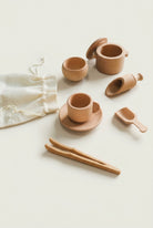 A collection of wooden sensory play tools including a pot with lid, cup, plate dismeter, bowl, scoops, tweezer, and two wooden sticks, displayed on a green surface with a drawstring bag.