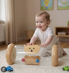A baby in a white onesie kneels on a white rug playing with a transparent plastic bear-shaped toy walker with tan wheels in a softly lit nursery.