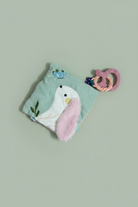 A baby teething toy set on a light wood surface, featuring a mint green corduroy pouch with a white bird applique, a fluffy pink ear, and a pink silicone teether ring.