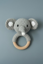 A handmade crocheted elephant rattle with a natural wooden ring handle, resting on a light-colored wooden surface.