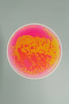 A circular liquid tile  with a vibrant abstract design featuring bright pink and yellow-orange paint splatters on a light gray background.