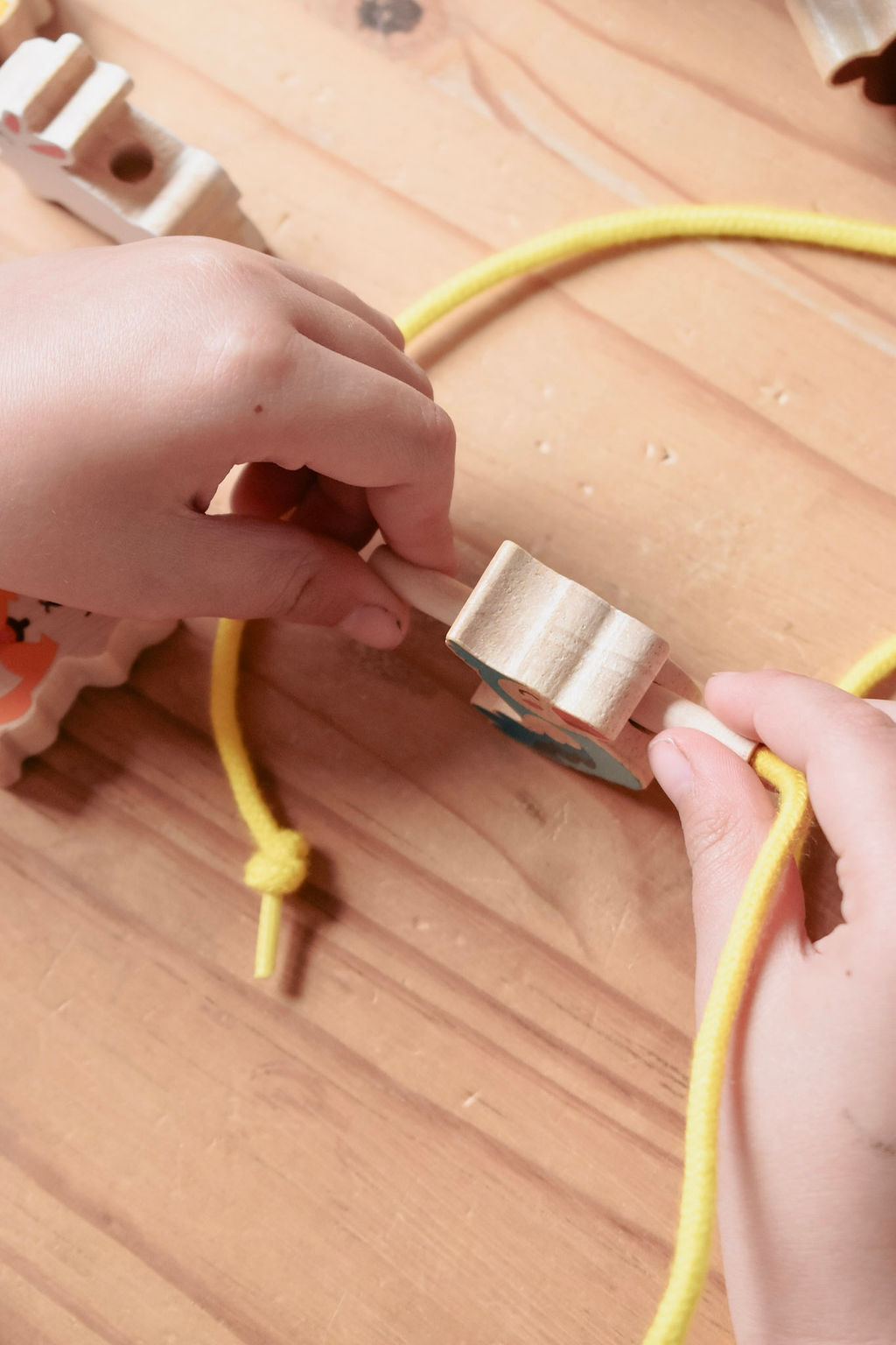 A close-up of hands tying a yellow rope around a small wooden bead on a light wood surface.