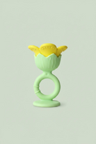 A baby teether toy with a light green silicone ring handle and a yellow flower-shaped silicone nibbler on top, resting on a light-colored wooden surface.