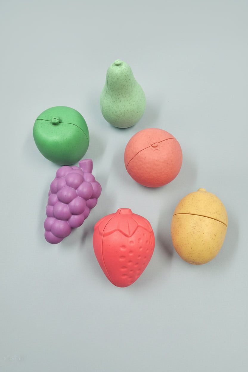 Six pastel fruit-shaped items arranged on a warm peach background: a green apple, a mint pear, a pink grapefruit, a purple grape cluster, a pink strawberry, and a pale yellow lemon. Each item has a split seam across the top half, suggesting they may be capsules, containers, or novelty fruit pieces.