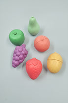 Six pastel fruit-shaped items arranged on a warm peach background: a green apple, a mint pear, a pink grapefruit, a purple grape cluster, a pink strawberry, and a pale yellow lemon. Each item has a split seam across the top half, suggesting they may be capsules, containers, or novelty fruit pieces.
