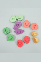 A set of colorful wooden shape-sorting blocks and matching cutters arranged on a soft peach background. The pieces are in pastel shades of green, pink, purple, and yellow, with different geometric and fruit-like shapes.