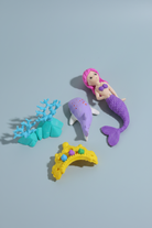A mermaid-themed toy set on a pink background, including a mermaid figure with a purple tail and pink hair, a purple dolphin, turquoise coral, a yellow arch bridge, and a small multicolored shell.