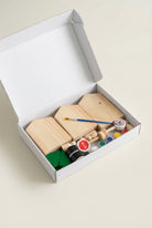 A wooden toy construction set inside an open white box, featuring light wood blocks, a small brush, colorful paint pots, and green tree pieces on a beige surface.