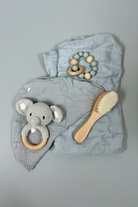 A baby gift set featuring a dusty blue muslin blanket set with a crocheted elephant teether, a wooden bead rattle, and a wooden baby brush, arranged on a light wood background.