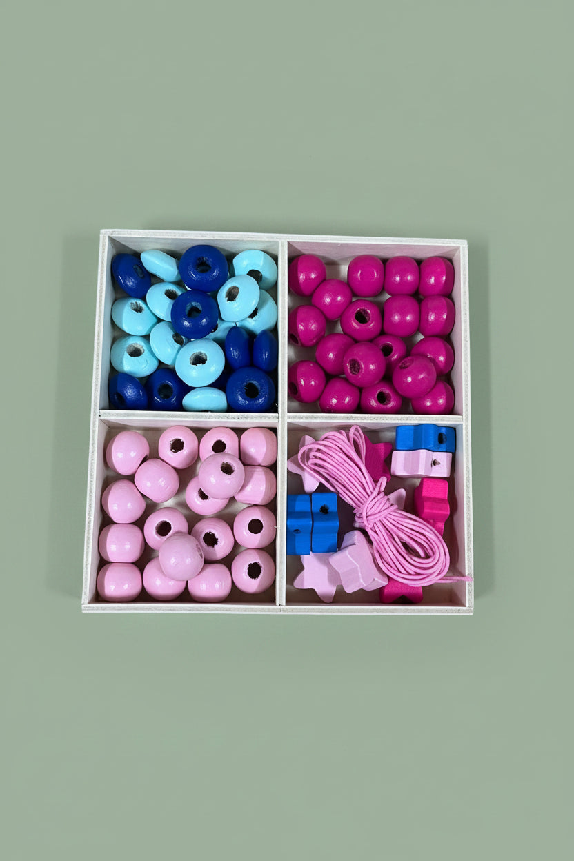A 4-compartment tray filled with colorful wooden beads and small wooden blocks, placed on a light wood surface. The beads are in pink, blue, and magenta, with some larger round beads and some small square blocks.