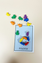 A set of small, colorful animal-shaped toy pieces arranged on a wooden surface around a printed card. The cards are white with black outlines and the brand name 'tiny tree toys' printed at the bottom.
