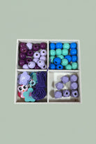 A 4-compartment wooden box filled with colorful wooden beads and small wooden charms, placed on a light wood tabletop. The beads come in purple, blue, mint green, and lavender colors.