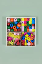 A wooden compartment box filled with colorful wooden beads and toy pieces, including round discs, square blocks, yellow cord, and ladybug-shaped beads, photographed on a light wood tabletop.