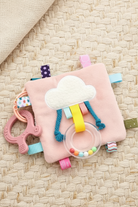 Soft square taggie comforter blanket in green and pink with a rainbow appliqué, colourful fabric tags around the edges, shown with a small ring with tiny balls rattle