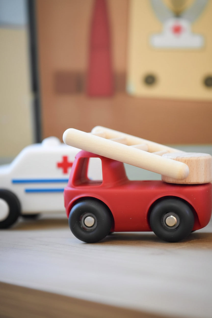 A wooden toy fire truck with a red body, black wheels, and a natural wood ladder on top, shown in a shallow depth-of-field close-up on a wooden surface.
