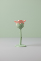 A small flower-shaped silicone teether with a pale green stem and base, and a pink flower head, placed on a light wood surface.