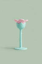 A pastel-colored flower-shaped silicone teether, featuring a light blue stem and base with a pink flower head, resting on a wooden surface.