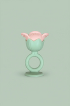 A baby teether toy with a mint green ring handle and a pink flower-shaped top, placed on a light wood surface.