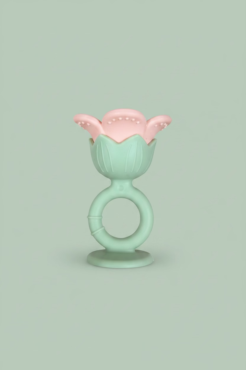 A baby teether toy with a mint green ring handle and a pink flower-shaped top, placed on a light wood surface.