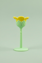 A small flower-silicone teether with a yellow top and a light green stem and base, photographed against a wooden surface.