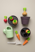A set of wooden toy gardening items including two pots with planters, a watering can, a trowel, carrots, radishes, and an onion, arranged on a light wood background.