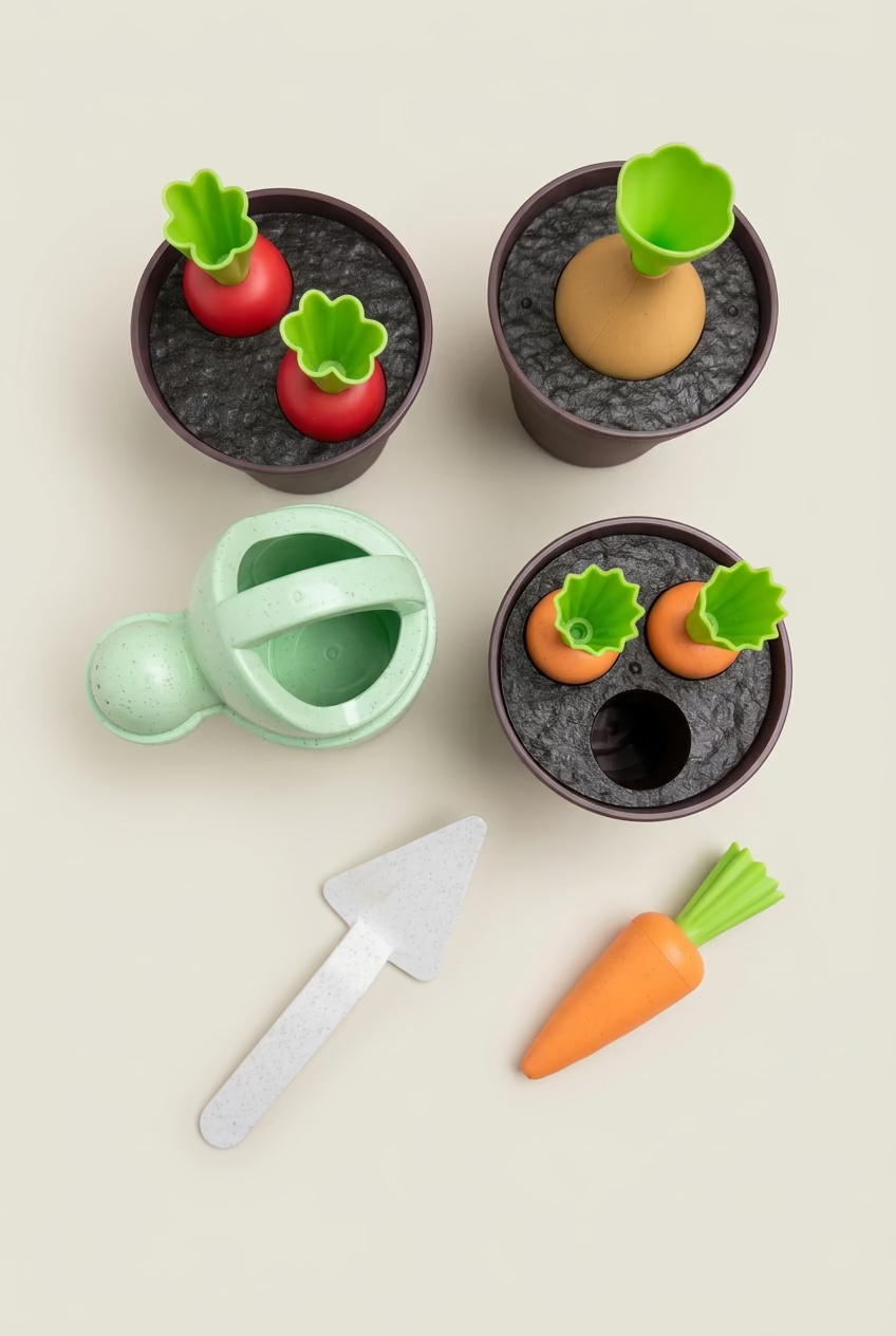 A children's wooden pretend-gardening set arranged on a light wood tabletop, including dark plant pots with toy vegetables, a mint green watering can, an orange carrot, and a silver-colored trowel.