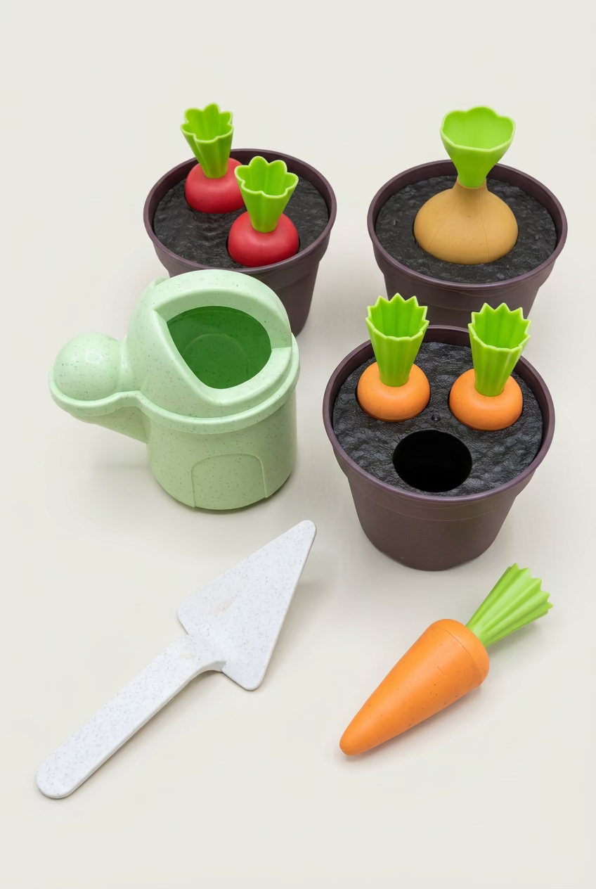 A children's toy gardening set on a wooden tabletop, including a light green watering can, dark brown pots with toy soil, orange carrots with green tops, red radishes, and a white trowel.