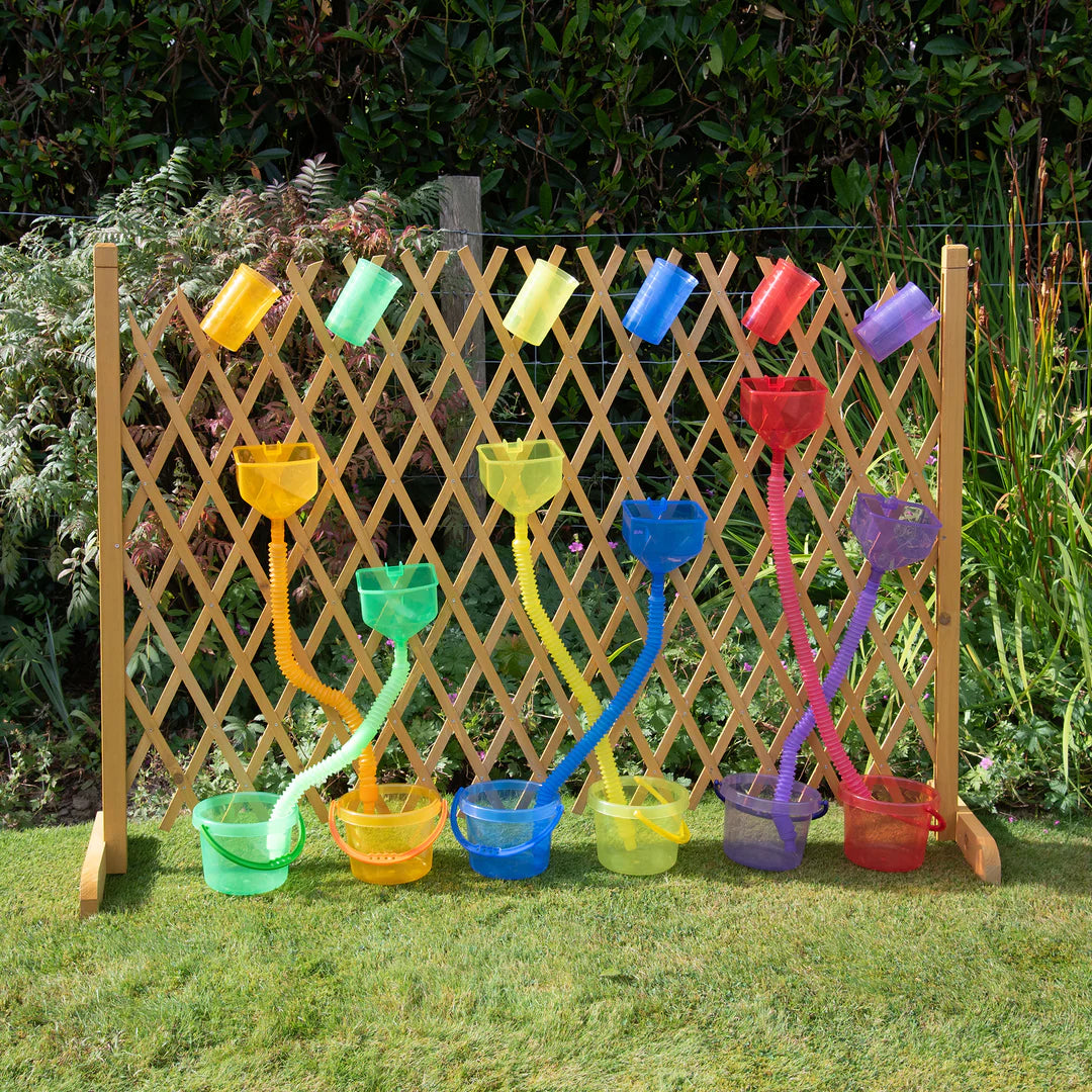 Colorful garden watering cans arranged against a wooden lattice fence with greenery.