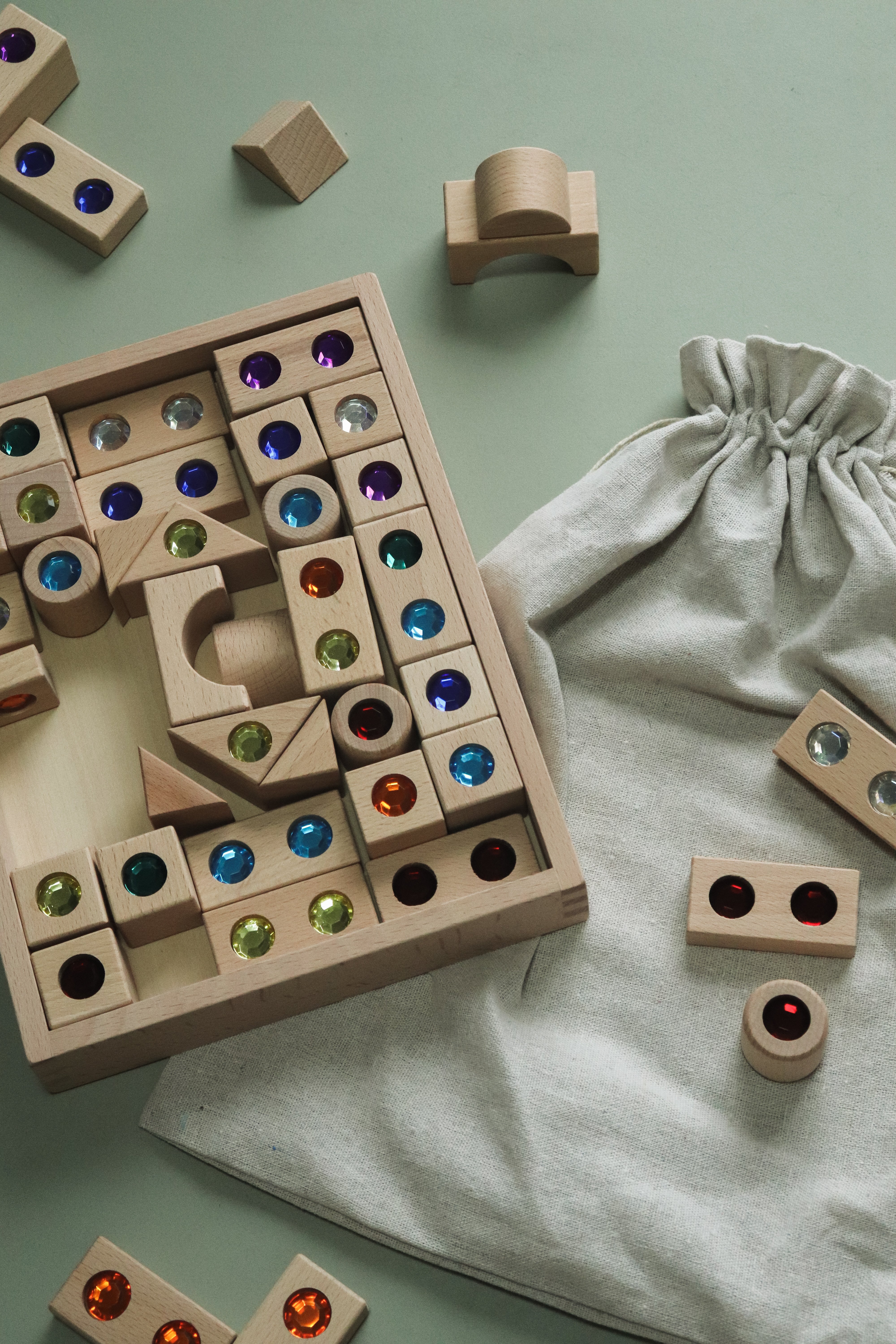 A wooden block puzzle game with colorful gemstone inserts, shown with a light gray fabric drawstring bag on a pale green background.