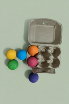 A cardboard egg carton with six colorful craft wooden balls in orange, pink, blue, green, yellow, and purple.