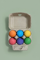 Six hand-painted wooden balls in orange, blue, yellow, pink, purple, and green, arranged inside an open brown cardboard egg carton on a light wood tabletop.