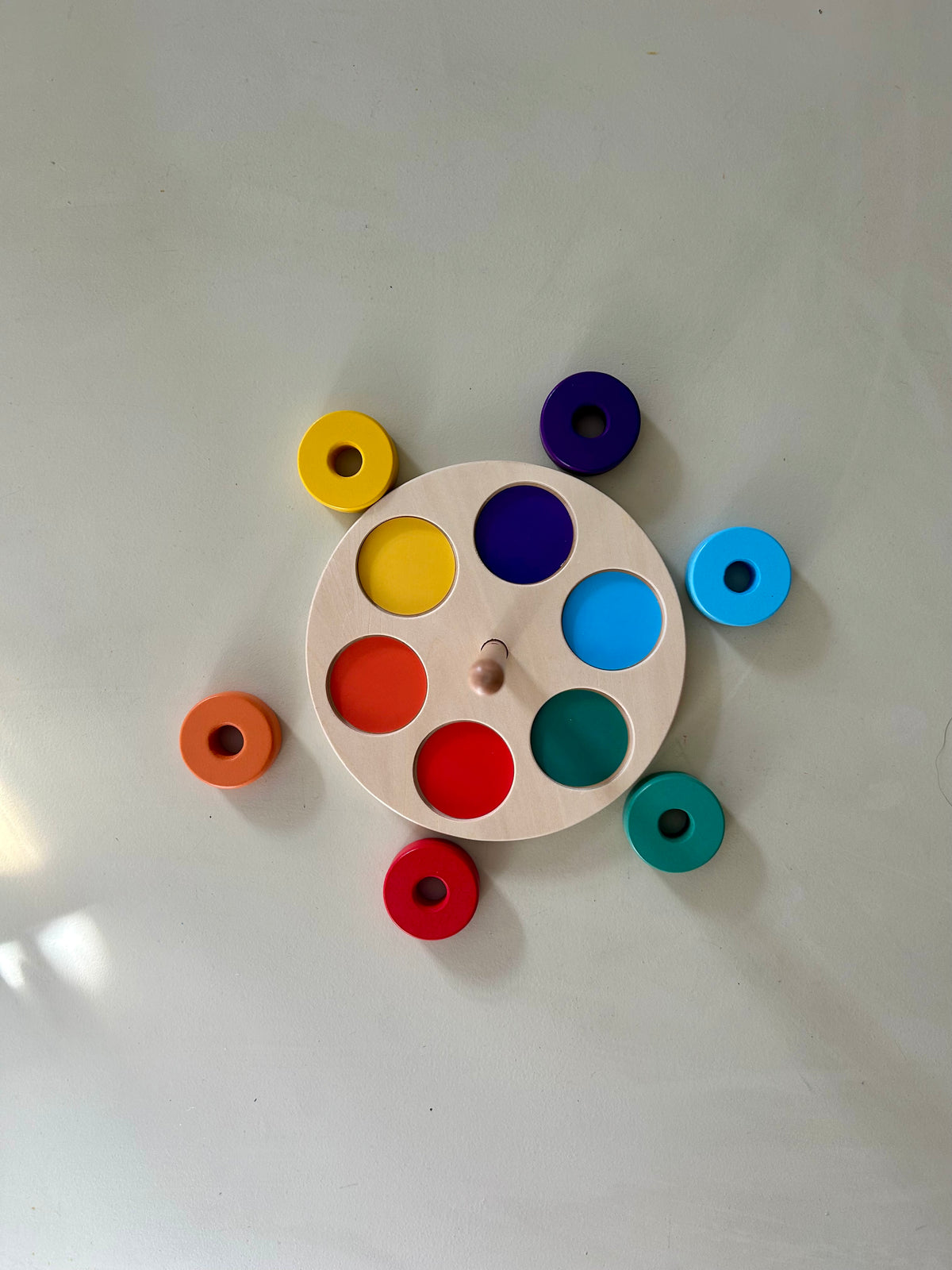 Overhead view of the round wooden base with six coloured circles, and six painted wooden rings laid out beside it in green, yellow, purple, red, orange and light blue.
