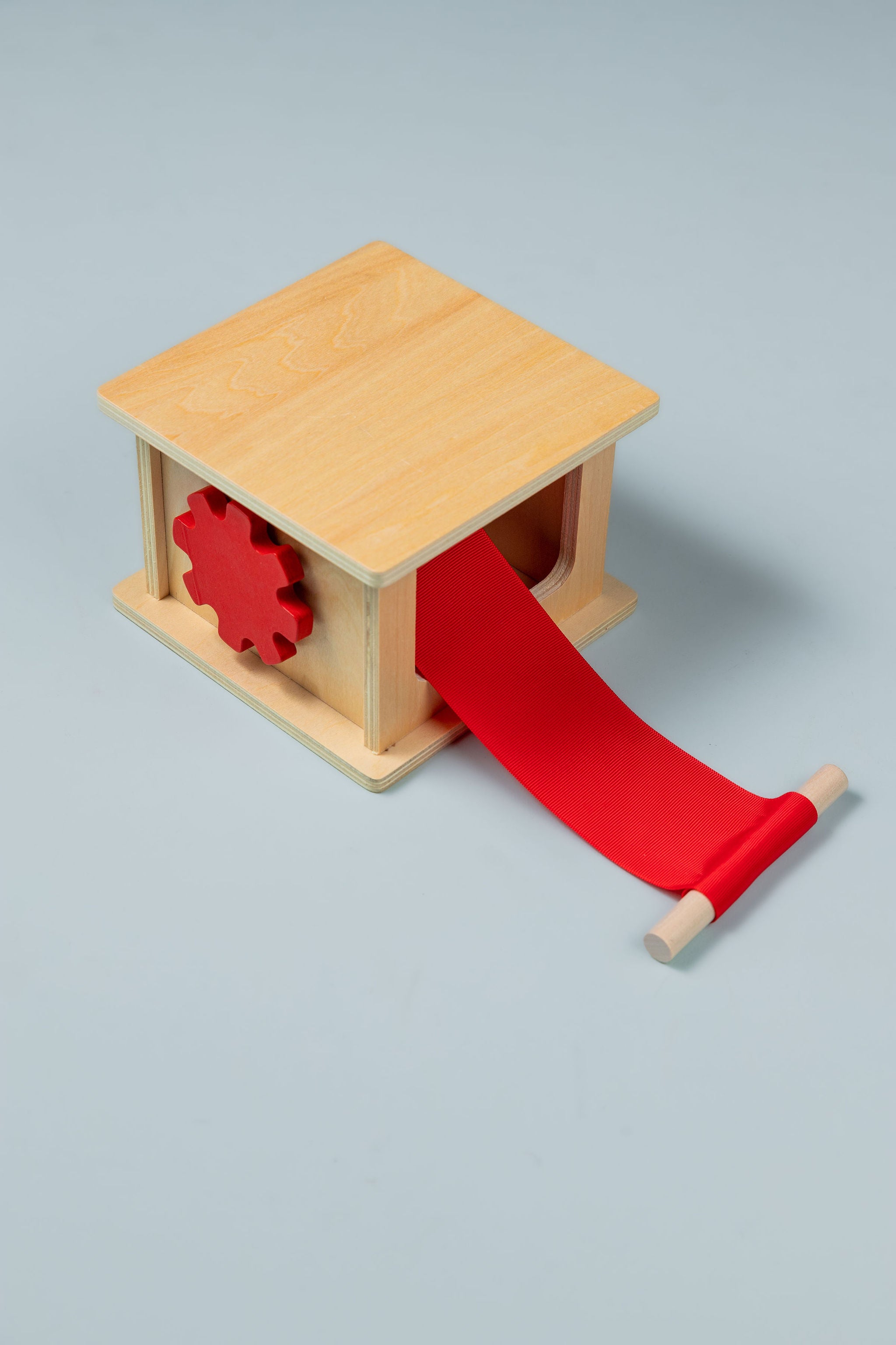 Natural wooden Montessori pulling and winding box with a red cog-shaped knob on the side and a red ribbon pulled out to a wooden dowel handle, on a pale blue-grey background.