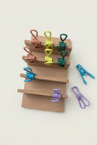 A set of colorful metal spring clips in multiple colors, including blue, purple, green, yellow, and pink, displayed on a small wooden stand with a few loose clips on a light wood tabletop.